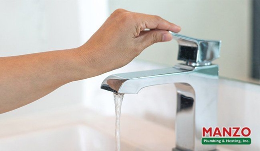 Close-up of a hand turning a chrome faucet on, water flowing into a sink; MANZO Plumbing & Heating logo.