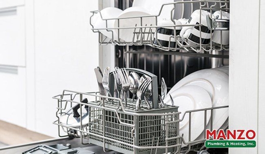 Open dishwasher with clean dishes and utensils in the racks; Manzo Plumbing logo visible in corner.