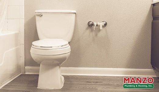 Residential bathroom with a white toilet, wall-mounted toilet paper holder, a bathtub to the left, and the Manzo Plumbing & Heating logo in the bottom-right corner.